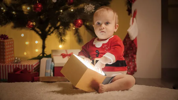 Menino em traje de Papai Noel sentado sob a árvore de Natal e segurando mágico brilhante presente de Natal em caixa de presente de ouro — Fotografia de Stock