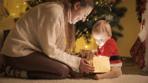 Счастливая улыбающаяся мать с маленьким сыном, сидящим рядом с елкой и открывающим коробку рождественского подарка — стоковое фото