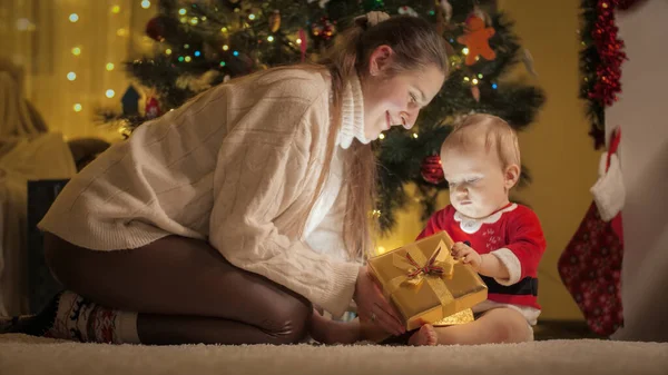 Mãe feliz com o bebê filho desembrulhando e olhando dentro da caixa de presente de Natal na sala de estar. Famílias e crianças comemorando as férias de inverno. — Fotografia de Stock