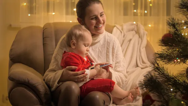 Glücklich lächelnde Mutter sitzt mit ihrem kleinen Sohn im Sessel neben dem leuchtenden Weihnachtsbaum. Familien und Kinder feiern Winterurlaub. — Stockfoto