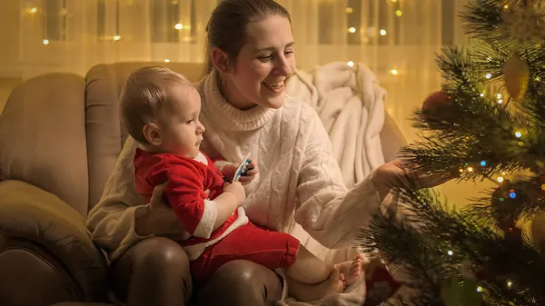 幼い男の子が若い母親とアームチェアに座って、輝く装飾されたクリスマスツリーを見ています。冬の休日を祝う家族や子供. — ストック写真
