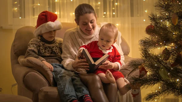 Glad leende mamma med barn läser sagobok i fåtölj bredvid julgran i vardagsrummet. Familjer och barn som firar vinterlov. — Stockfoto