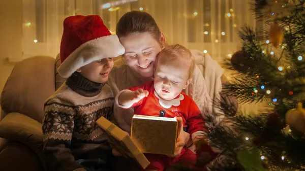 Glad familj med barn som sitter i fåtölj vid julgranen och tittar inuti glödande julklappslåda. Familjer och barn som firar vinterlov. — Stockfoto