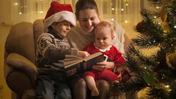 Niño con hermano y madre leyendo un libro de cuentos de hadas en Nochebuena. Familias y niños celebrando las vacaciones de invierno. —  Fotos de Stock