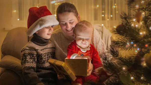 Netter kleiner Junge sitzt auf dem Schoß der Mutter im Sessel und schaut in die leuchtende Weihnachtsgeschenkbox. Familien und Kinder feiern Winterurlaub. — Stockfoto