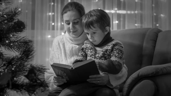 Svart och vit bild av glad leende pojke lyssnar på sagobok på julafton. Familjer och barn som firar vinterlov. — Stockfoto