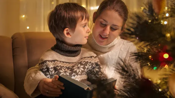 Mladá matka se synem slaví Štědrý večer a čtení pohádkové knihy v křesle. Rodiny a děti slaví zimní prázdniny. — Stock fotografie