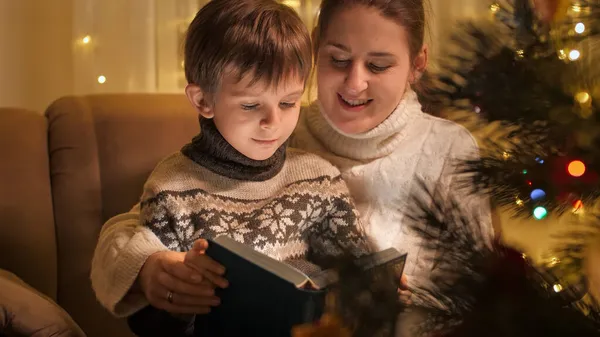 Glad leende pojke med mor läser sagobok bredvid glödande dekorerad julgran. Familjer och barn som firar vinterlov. — Stockfoto