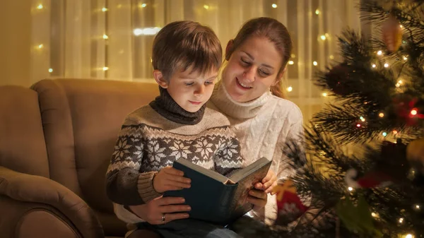 Portrait of smiling mother with son reading Christmas story book next to glowing Christmas tree. Families and children celebrating winter holidays. — Stock Photo, Image