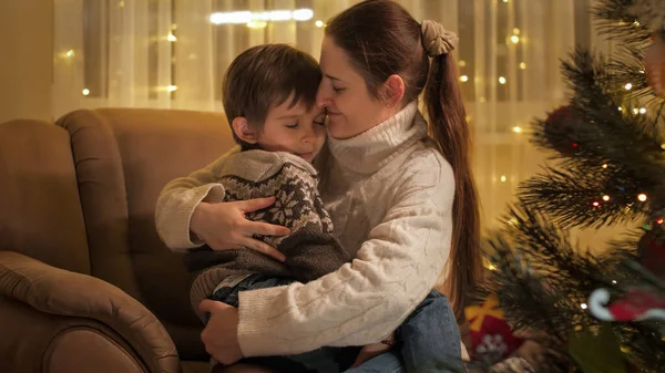 Rapaz giro a abraçar a mãe na poltrona ao lado da árvore de Natal. Famílias e crianças comemorando as férias de inverno. — Fotografia de Stock
