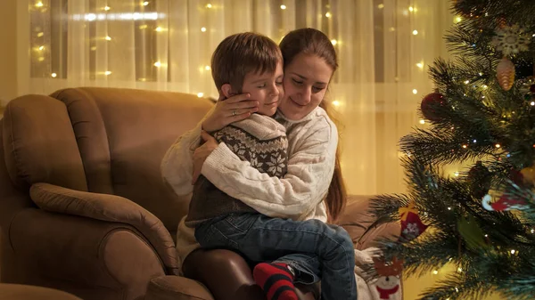 Mãe e filho felizes sentados em poltrona ao lado da árvore de Natal e abraços. Famílias e crianças comemorando as férias de inverno. — Fotografia de Stock