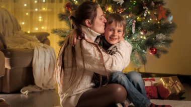 Olumlu gülümseyen anne ve oğul Noel arifesinde oturma odasında sarılıp öpüşüyorlar. Aileler ve çocuklar kış tatilini kutluyor.