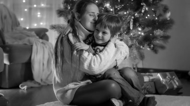 Mutlu gülümseyen anne ve oğlunun siyah beyaz portresi. Evde parlayan Noel ağacının yanında sarılıyor.