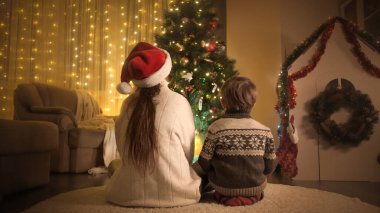 Oturma odasındaki Noel ağacının altında sarılan anne ve oğlanın arka görüntüsü. Aileler ve çocuklar kış tatilini kutluyor.