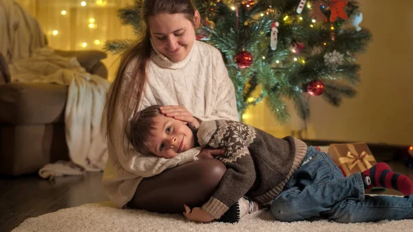 輝くクリスマスツリーの横に横たわっている彼女の笑顔の息子を愛撫若い母親。冬の休日を祝う家族や子供. — ストック写真