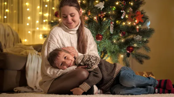 Счастливый улыбающийся мальчик лежит на коленях у матерей, пока она ласкает его рядом с ёлкой. Семьи и дети празднуют зимние праздники. — стоковое фото