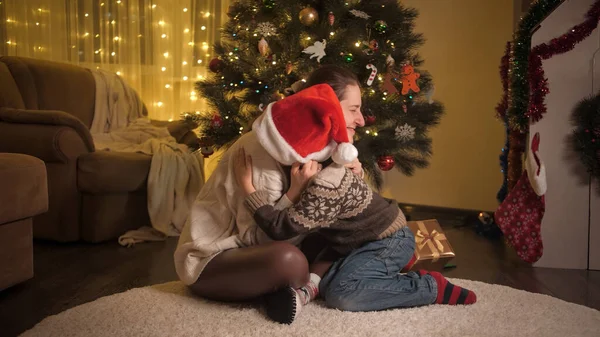 Jovem mãe colocando chapéu de Papai Noel em seu filho e abraçando-o sob a árvore de Natal. Famílias e crianças comemorando as férias de inverno. — Fotografia de Stock