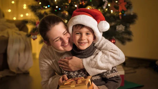 Улыбающаяся мать дарит рождественский подарок в золотой коробке своему улыбающемуся мальчику в шляпе Санты. Семьи и дети празднуют зимние праздники. — стоковое фото