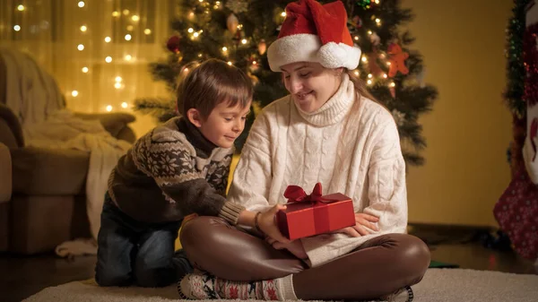 Glada leende pojke med mamma ger julklappar och kramar under julgranen hemma. Familjer och barn som firar vinterlov. — Stockfoto