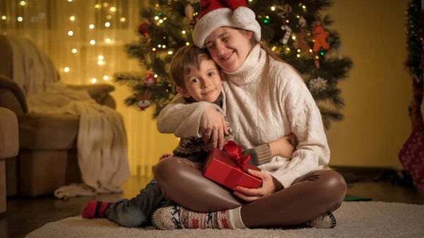 A kisfiú karácsonyi meglepetést csinál, és ajándékot ad az anyjának Mikulás sapkában. A téli ünnepeket ünneplő családok és gyermekek. — Stock Fotó