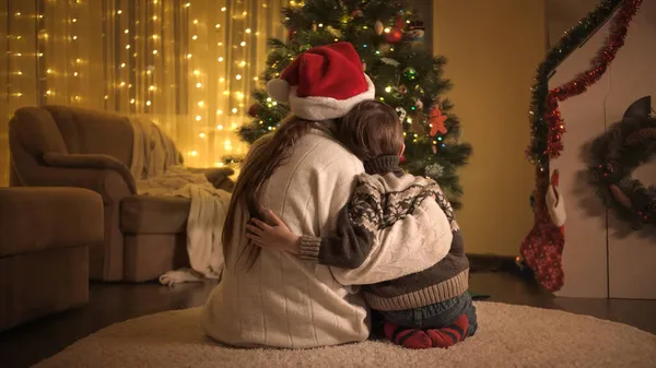 Visão traseira da mãe em chapéu de Santa e filho em suéter de malha sentado junto à árvore de Natal e olhando para guirlanda de luz brilhante — Fotografia de Stock