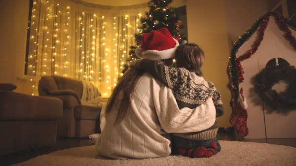 Jovem mãe abraçando seu filhinho e olhando para a árvore de Natal brilhante na sala de estar. Famílias e crianças comemorando as férias de inverno. — Fotografia de Stock