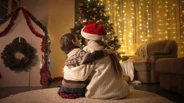 小さな男の子とお母さんで暖かいウールセーターに座って隣に美しい輝くクリスマスツリーとお祝い新年に自宅 — ストック写真