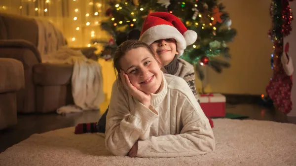Fröhlicher Junge liegt auf Mutter neben leuchtend geschmücktem Weihnachtsbaum. Familien und Kinder feiern Winterurlaub. — Stockfoto