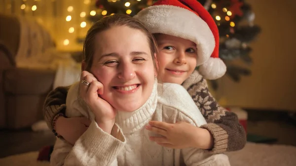 Nahaufnahme Porträt eines kleinen Jungen mit Weihnachtsmütze, der seine Mutter neben dem Weihnachtsbaum umarmt. Pure Emotionen bei Familien und Kindern, die Winterurlaub feiern. — Stockfoto