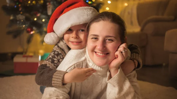 Porträt einer lächelnden Mutter mit Sohn in Nikolausmütze, die an Heiligabend auf dem Boden liegt. Pure Emotionen bei Familien und Kindern, die Winterurlaub feiern. — Stockfoto