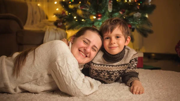 Portret uśmiechniętej mamy z synem obejmującym i leżącym na dywanie pod choinką w salonie. Czyste emocje rodzin i dzieci świętujących zimowe wakacje. — Zdjęcie stockowe