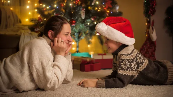 Garoto sorridente com a mãe deitada cara a cara no tapete sob a árvore de Natal e pálido com chapéu de Papai Noel. Emoções puras de famílias e crianças que celebram férias de inverno. — Fotografia de Stock
