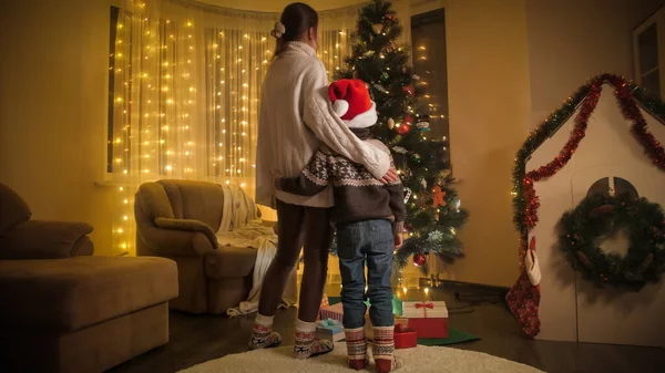 サンタの帽子の息子と母はリビングルームに立って、カラフルな輝くクリスマスツリーを見ています。冬休みを祝う家族や子供たちの純粋な感情. — ストック写真