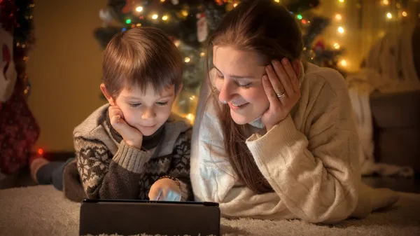 Mãe sorridente com o filho pequeno usando computador tablet sob a árvore de Natal em casa. Emoções puras de famílias e crianças que celebram férias de inverno. — Fotografia de Stock