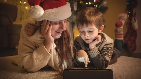 Matka s dítětem na Štědrý večer pomocí digitálního tabletu počítače vedle vánočního stromečku. Čisté emoce rodin a dětí oslavujících zimní prázdniny. — Stock fotografie