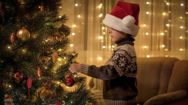 Retrato de menino pendurado bable vermelho em brilhante árvore de Natal colorido na sala de estar. Emoções puras de famílias e crianças que celebram férias de inverno. — Fotografia de Stock