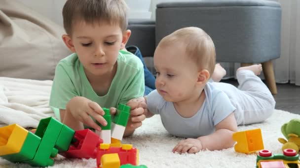 Niño pequeño con el hermano pequeño construyendo torre de bloques de juguete y ladrillos en el suelo en la sala de estar. Concepto de desarrollo infantil, educación y creatividad en el hogar — Vídeos de Stock
