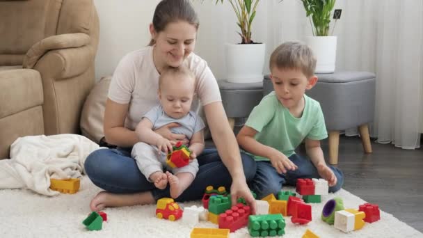 Маленький мальчик и старший брат играют с мамой в игрушки на ковре в гостиной. Концепция семейного времяпровождения и развития детей — стоковое видео