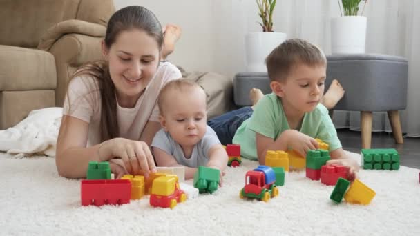 Χαμογελώντας χαρούμενα μωρά, αγόρι και μητέρα χαζολογούν παιχνίδια στο χαλί στο σπίτι. Έννοια της οικογένειας έχοντας χρόνο μαζί και την ανάπτυξη των παιδιών — Αρχείο Βίντεο