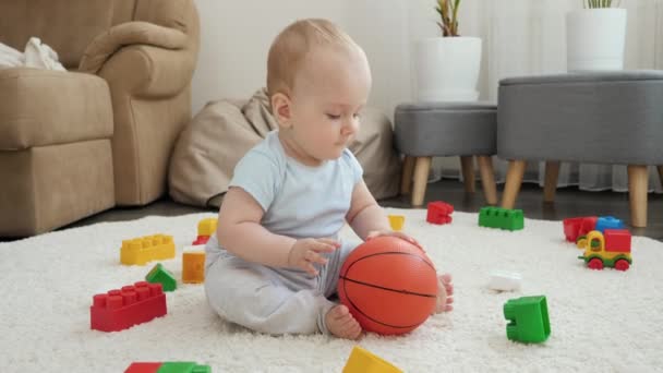Feliz niño sonriente sentado en la alfombra y jugando con la pelota de baloncesto en casa. Concepto de desarrollo infantil, deportes, educación y creatividad en el hogar — Vídeos de Stock