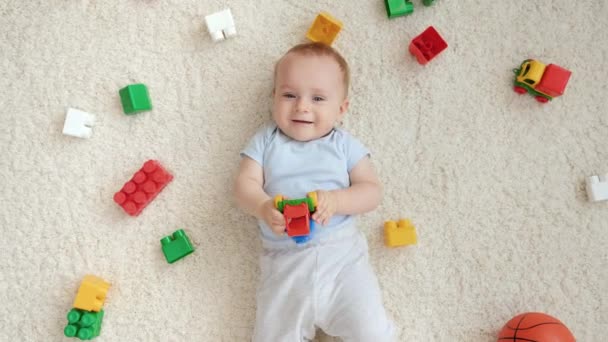 Feliz niño sonriente con un montón de juguetes tumbados en la alfombra y mirando hacia arriba en la cámara. Concepto de desarrollo infantil, educación y creatividad en el hogar — Vídeos de Stock