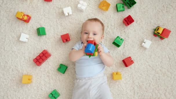 Portret van een lachend jongetje omringd door veel speelgoed liggend op tapijt in de speelkamer. Begrip "ontwikkeling, onderwijs en creativiteit van kinderen thuis" — Stockvideo