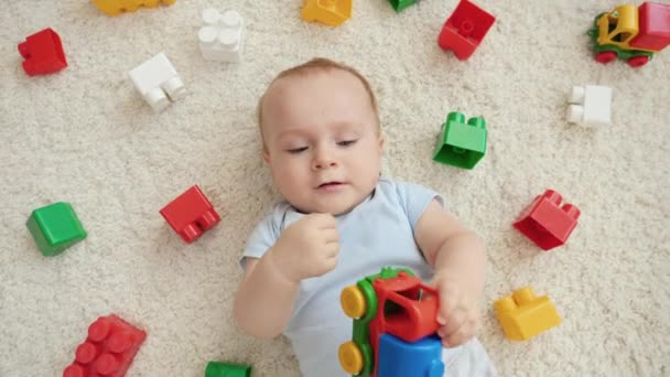 Lindo niño jugando coche de juguete en la alfombra junto a un montón de juguetes colroful, bloques y ladrillos. Concepto de desarrollo infantil, educación y creatividad en el hogar — Vídeos de Stock