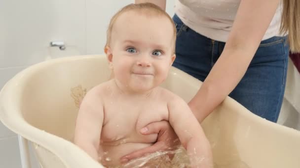 Szczęśliwy, śmiejący się chłopczyk siedzący w plastikowej wannie i rozpryskujący wodę. Pojęcie higieny dzieci, opieki zdrowotnej i rodzicielstwa. — Wideo stockowe
