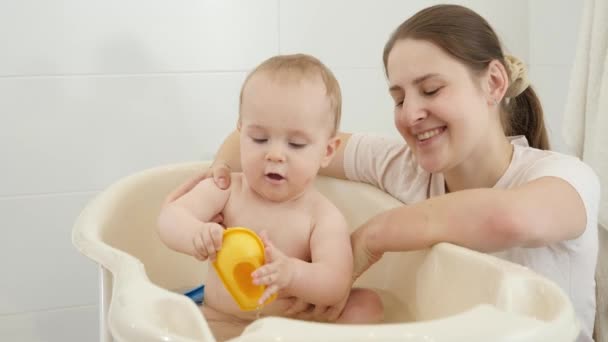Feliz madre sonriente mirando a su pequeño hijo salpicando agua y jugando en el baño. Concepto de higiene infantil, atención sanitaria y crianza de los hijos. — Vídeos de Stock