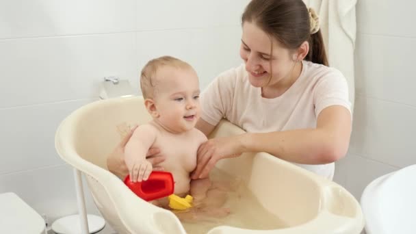 Madre sonriente sosteniendo a su pequeño hijo jugando con botes de juguete en el baño. Concepto de higiene infantil, atención sanitaria y crianza de los hijos. — Vídeos de Stock