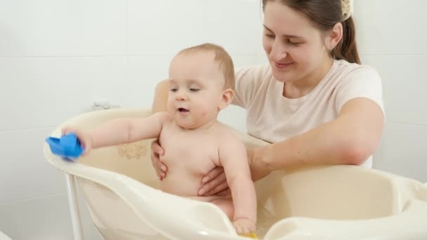 Glimlachende moeder die vasthoudt en haar zoontje thuis in bad ziet spelen. Concept van kinderhygiëne, gezondheidszorg en ouderschap. — Stockvideo