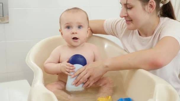 かわいい赤ちゃんの男の子が座って、自宅で小さなプラスチック製のバスルームで洗濯します。子供の衛生、医療、子育ての概念. — ストック動画