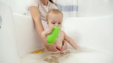 Gülümseyen çocuk köpükle banyo yapıyor ve oyuncak teknelerle oynuyor. Evde çocuk hijyeni, sağlık ve geliştirme kavramı