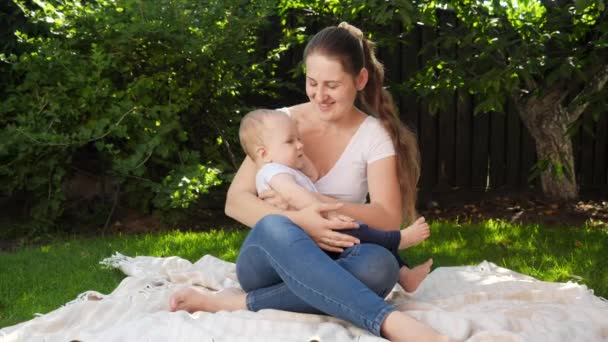 Boldog mosolygó anya ringatja a kisfiát, miközben a kertben ül a takarón. Szülői, család, gyermekek fejlődését, és szórakoztató szabadban a természetben. — Stock videók